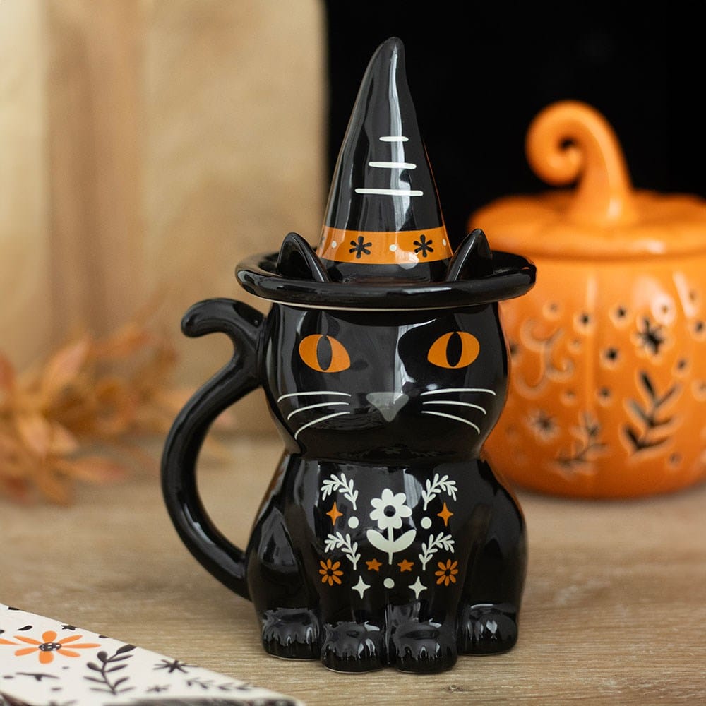 Frightful Folk witchy black cat Halloween lidded mug with pointed hat, orange eyes and floral details beside an orange pumpkin decoration
