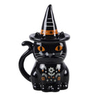 Front view of witchy black cat Halloween lidded mug with orange eyes, floral folk artwork, cat-tail handle and pointed witch's hat