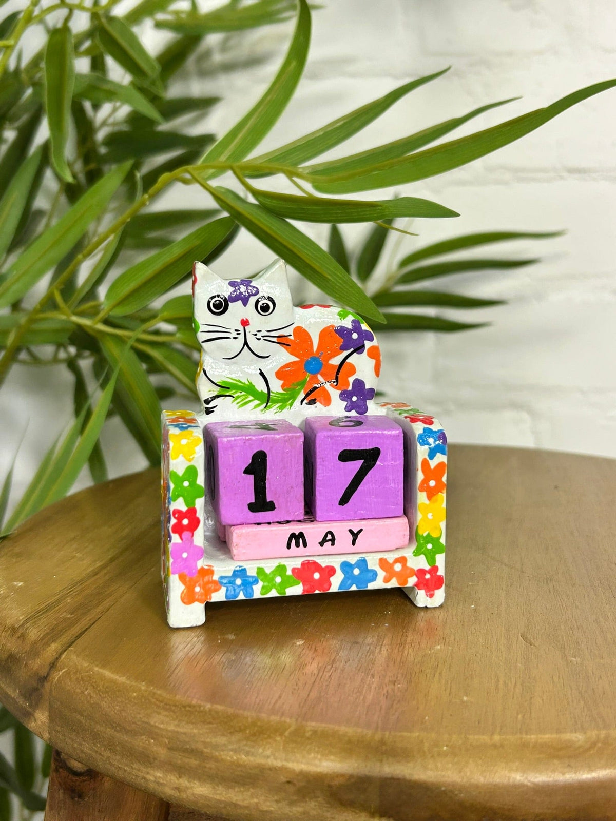 Colourful hand-painted wooden cat perpetual calendar displayed on a wooden table, with purple date blocks and floral decorations.
