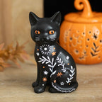 Frightful Folk black cat Halloween ornament with orange eyes, white botanical markings and stars displayed beside an orange pumpkin decoration