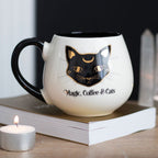 Magic, Coffee & Cats rounded ceramic mug with black cat design displayed on a book beside a candle.