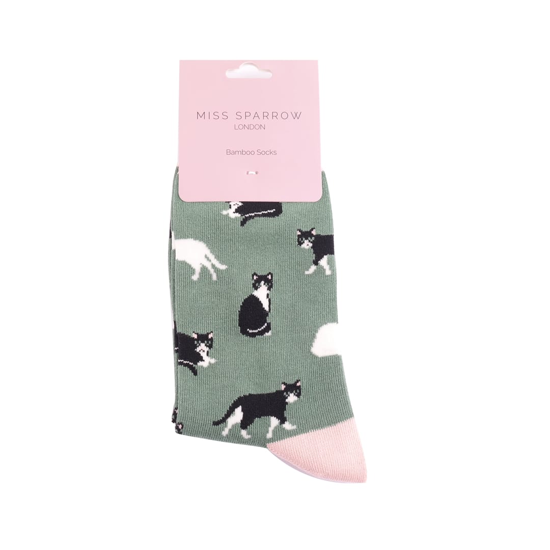 Pair of Miss Sparrow Ladies Bamboo Socks in retail packaging in soft green featuring playful black and white cat silhouettes on a sage green background with pale pink heels, toes and cuffs, made from a soft bamboo-rich fabric. 