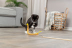 Black and white kitten playing with a yellow Trixie Plush Cat Teaser on a wooden floor.