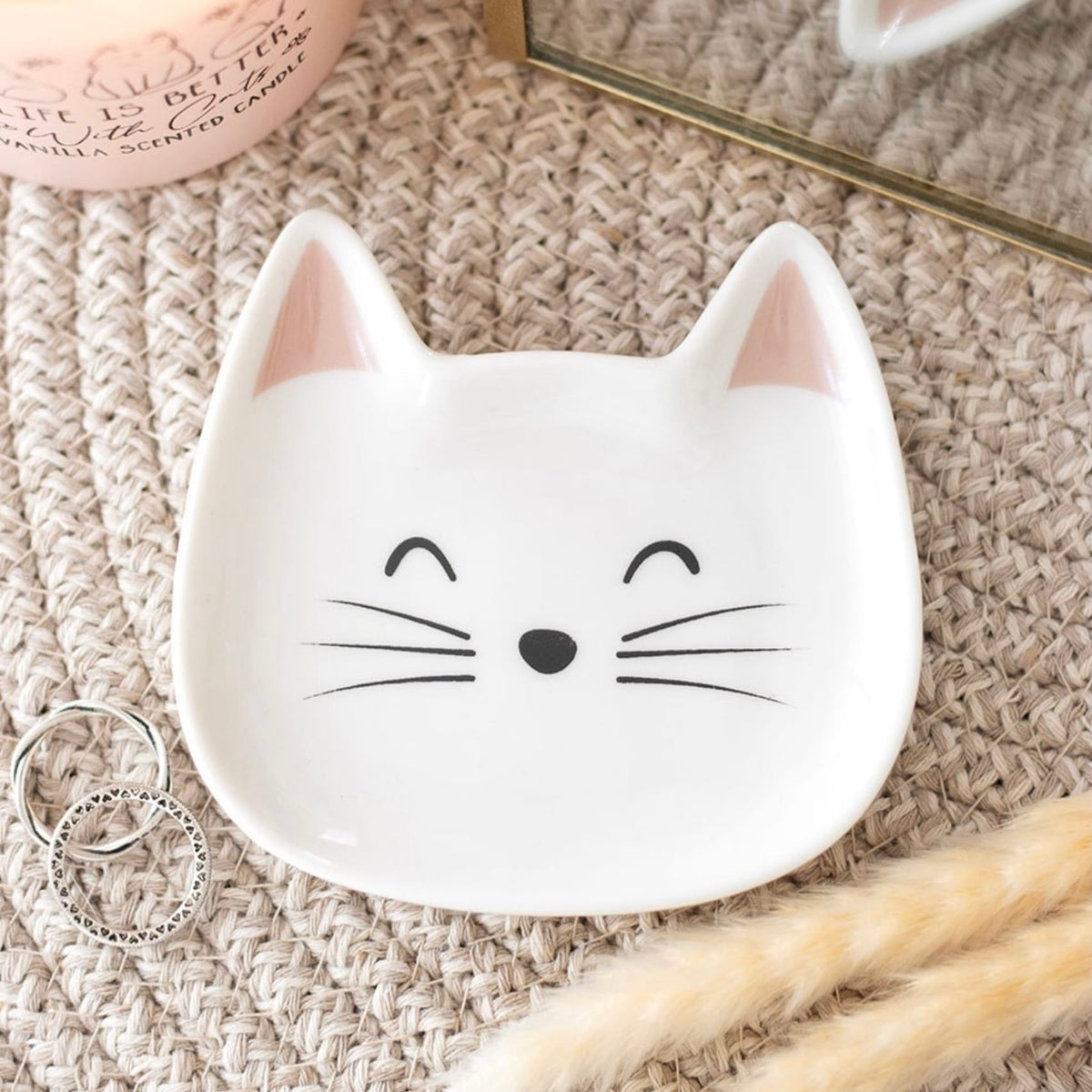 White cat face ceramic jewellery dish holding silver rings on a textured dressing table beside a candle, showing its use as a jewellery holder