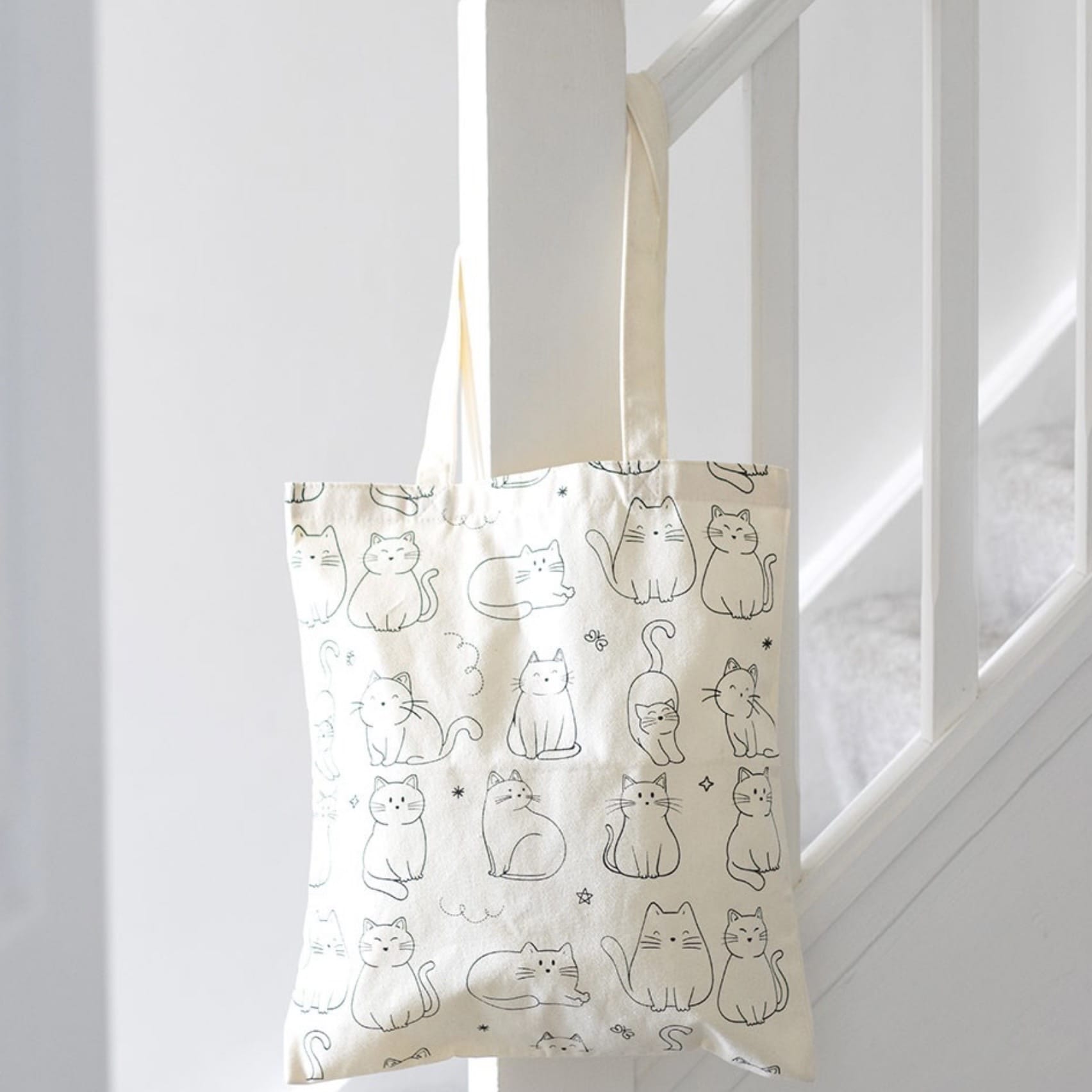 White cat print polycotton tote shopping bag hanging from a stair rail, showing its long handles and playful feline design