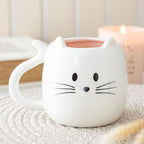 Cute white ceramic cat mug with pink interior, pointed ears, whiskered face and curled tail handle on a cosy tabletop.